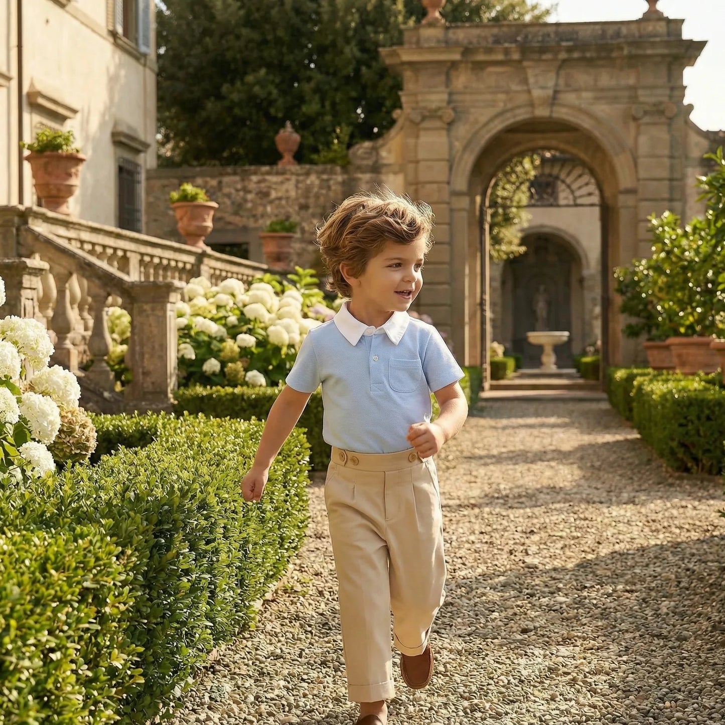 Boy wearing Maison Leopolda Henry Trousers in beige and Oscar Polo Shirt in light blue walking through an Italian villa garden with white hydrangeas and classical stone archway