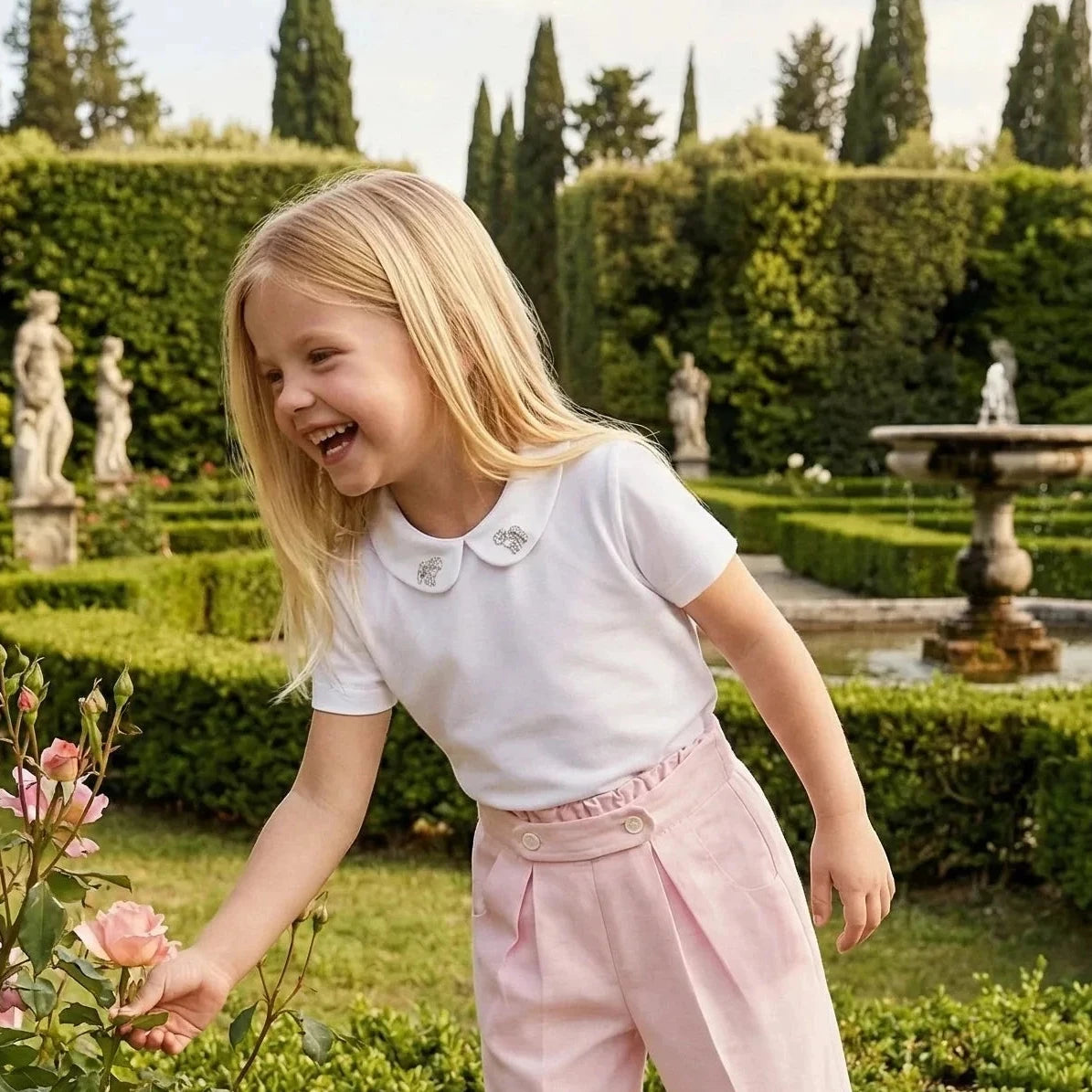 Girl wearing Maison Leopolda Léonie Trousers in pink and Rose Blouse in white laughing in an Italian garden with manicured hedges and fountain