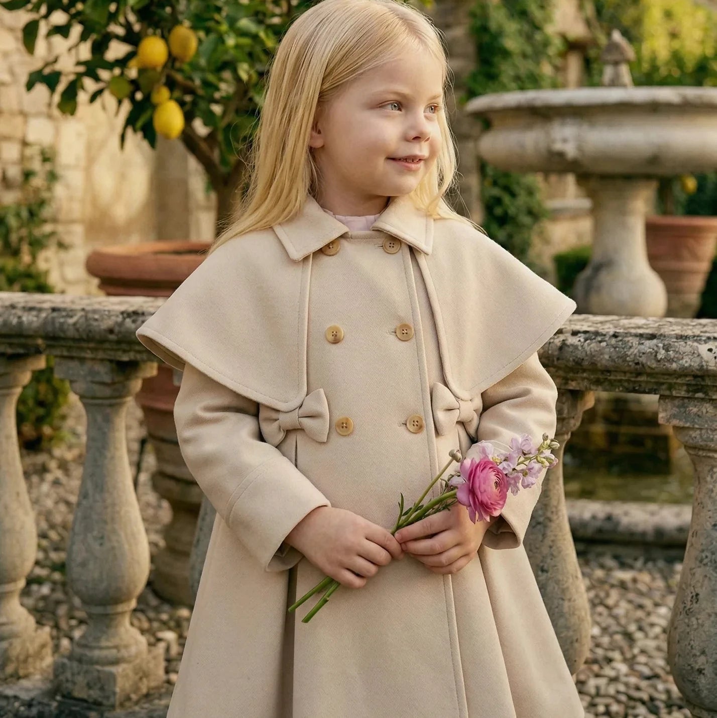 Girl wearing Maison Leopolda Alba Trench-Coat in beige holding flowers by a fountain in an Italian garden