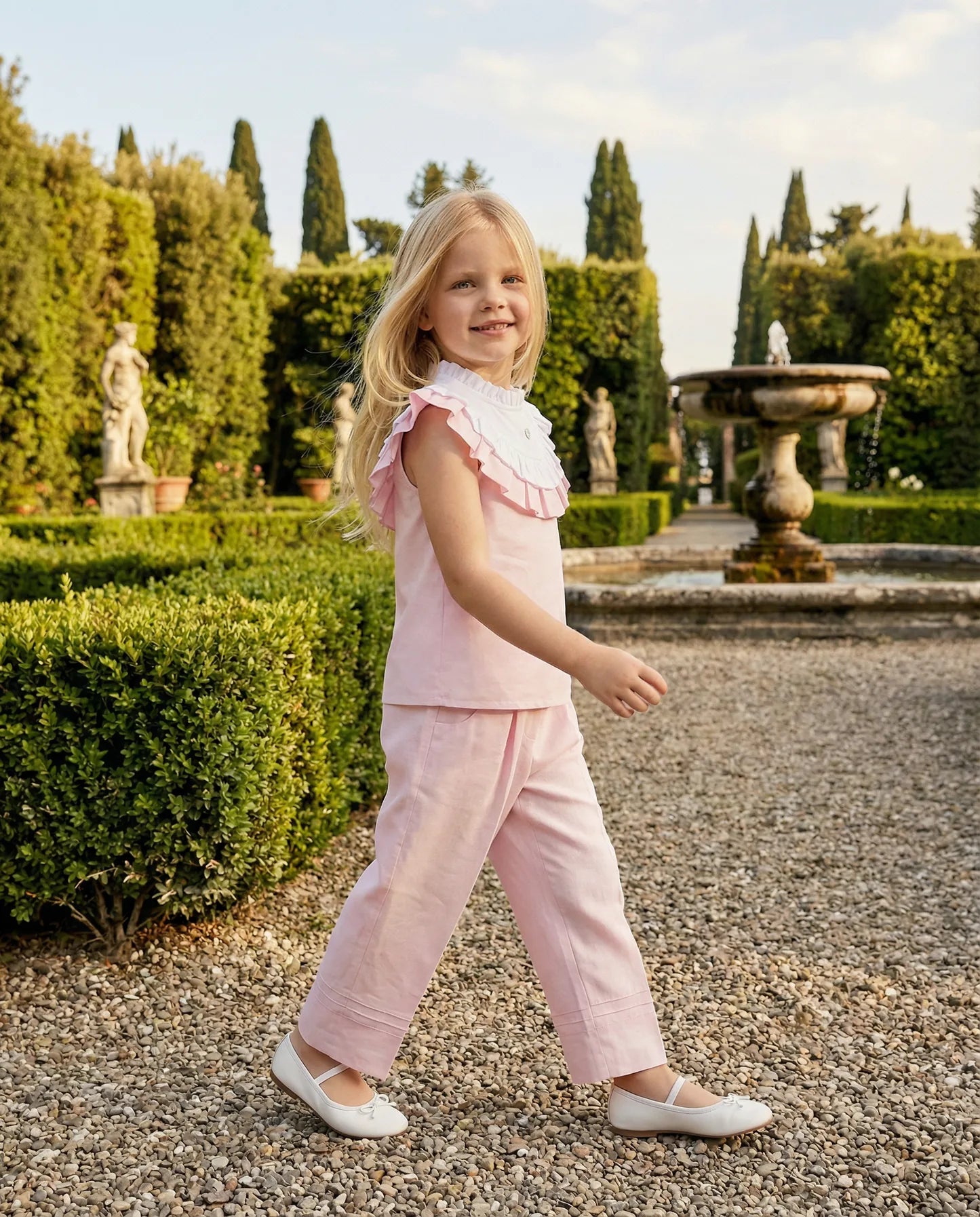 Girl wearing the pink Rose blouse and Léonie trousers walking through an Italian garden