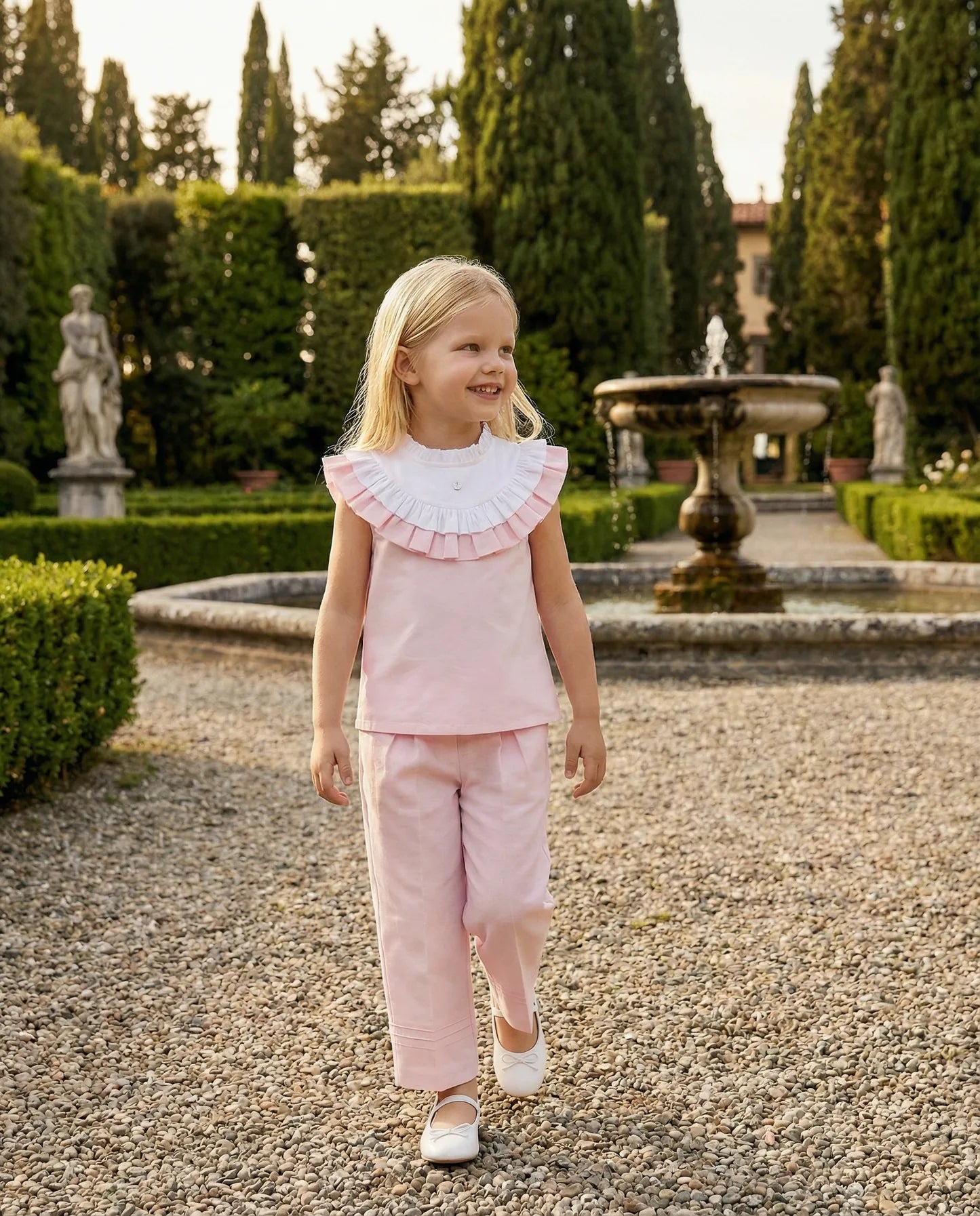 Girl in the pink Rose blouse and Léonie trousers on a gravel path by a garden fountain